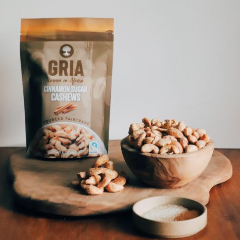 A bag of GRIA’s Fairtrade Cinnamon Sugar Cashews sits atop a wooden board. To the right of the bag is a wooden bowl displaying those cashews. Alongside it is a shallow bowl with cinnamon sugar.