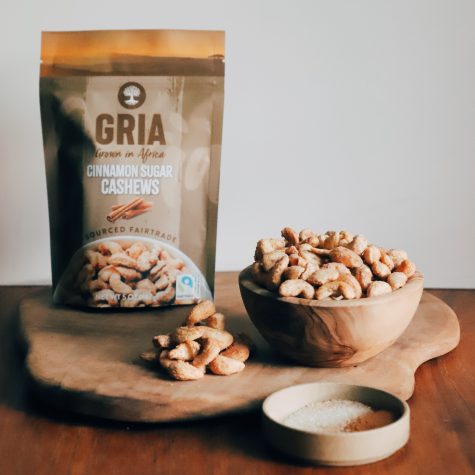 A bag of GRIA’s Fairtrade Cinnamon Sugar Cashews sits atop a wooden board. To the right of the bag is a wooden bowl displaying those cashews. Alongside it is a shallow bowl with cinnamon sugar.