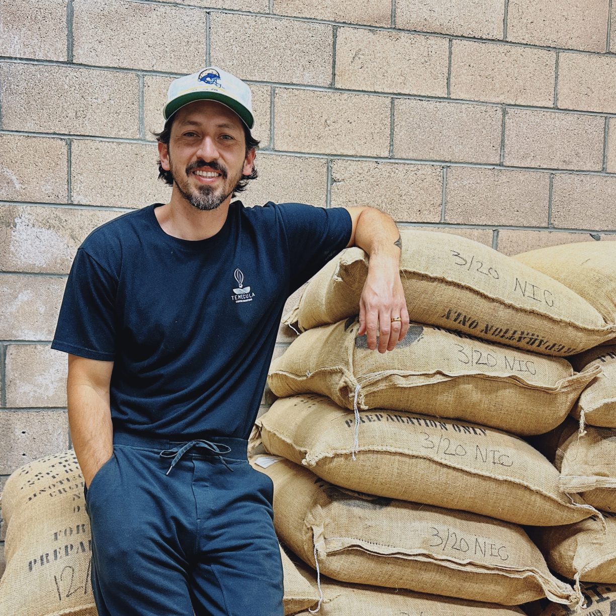 Ed Burga leans against a stack of sacks containing coffee beans. His shirt has the Temecula Coffee Roasters logo on it.