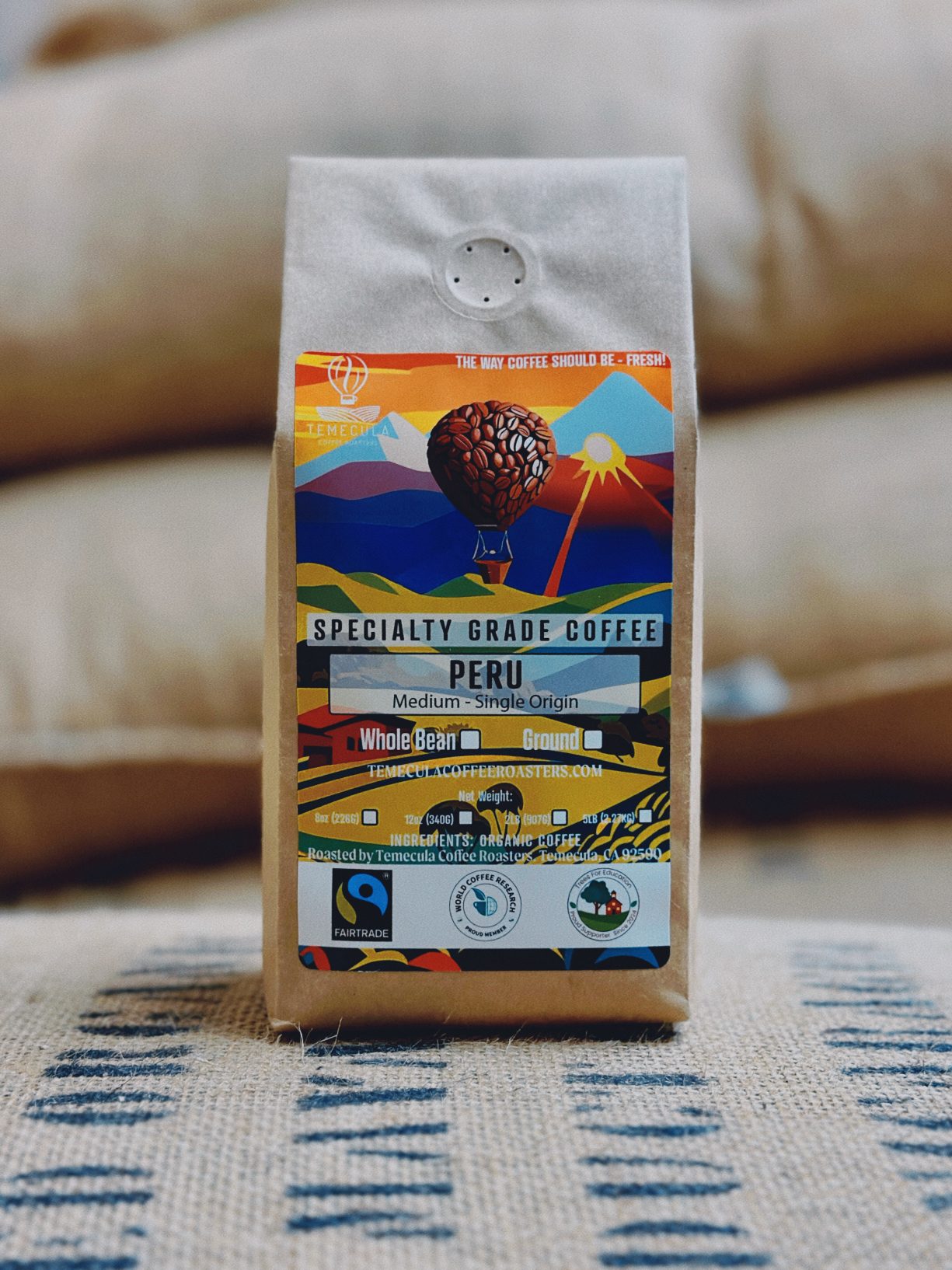 A bag of Temecula Coffee Roaster's Fairtrade, specialty grade coffee from Peru sits atop a stack of sacks containing coffee beans.