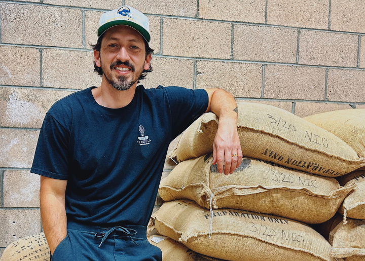 Ed Burga leans against a stack of sacks containing coffee beans. His shirt has the Temecula Coffee Roasters logo on it.