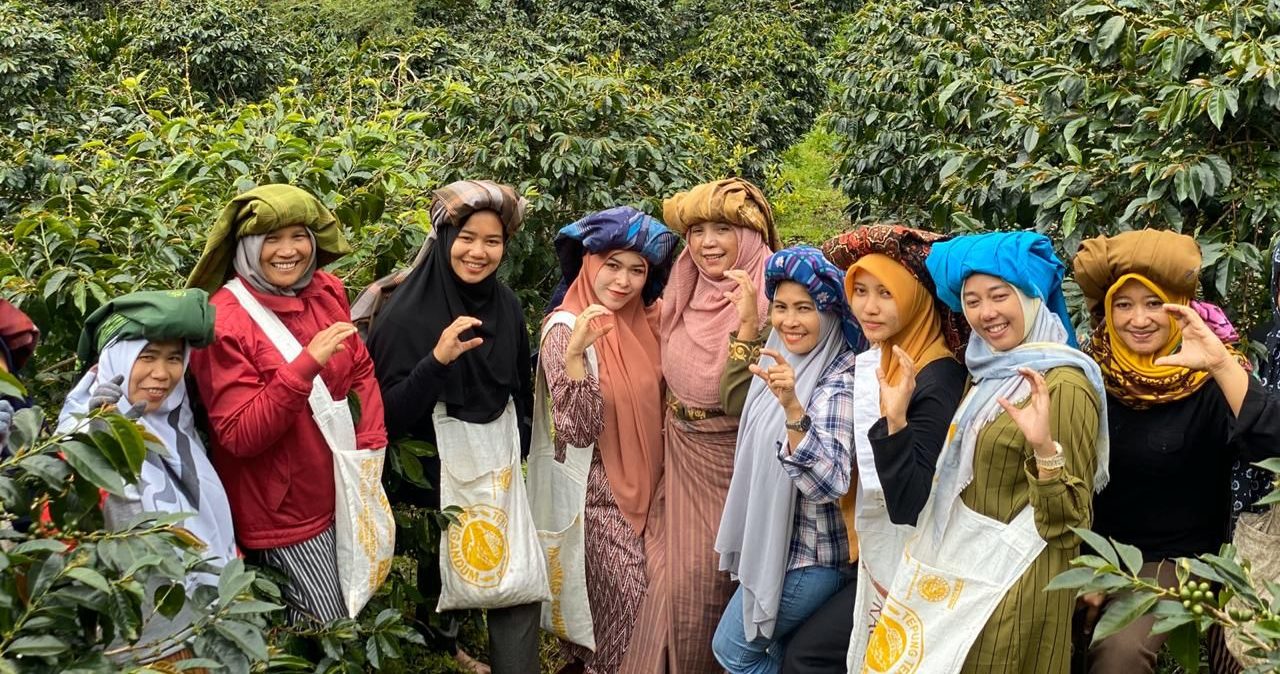 A group of women farmers pose amid a coffee farm. They shape their hands into the letter 