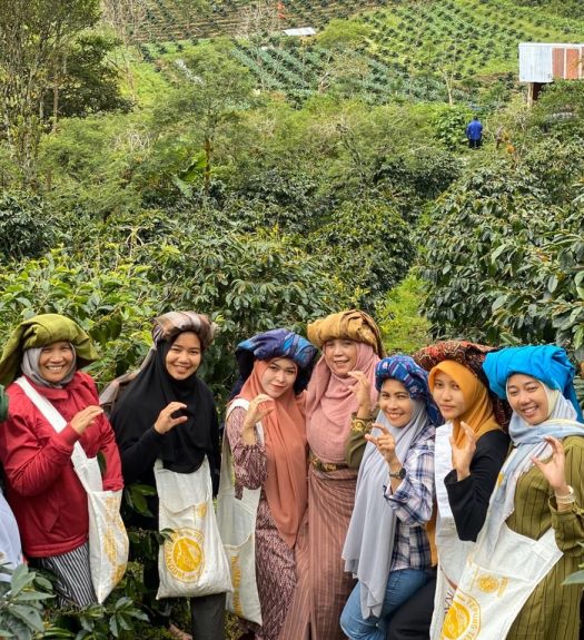 A group of women farmers pose amid a coffee farm. They shape their hands into the letter 