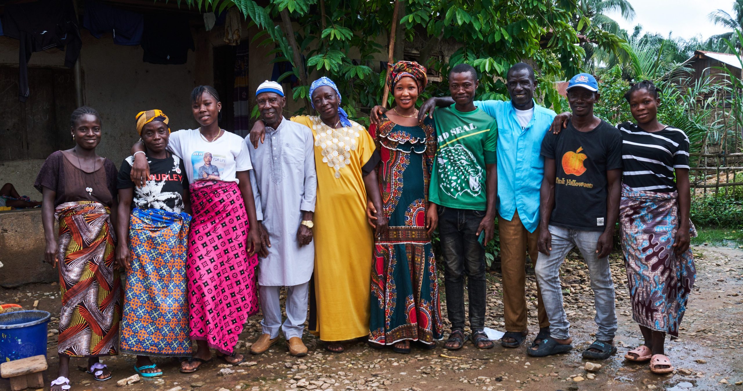 A group portrait of Munafa cocoa farmers standing outside on packed soil in front of green foliage.