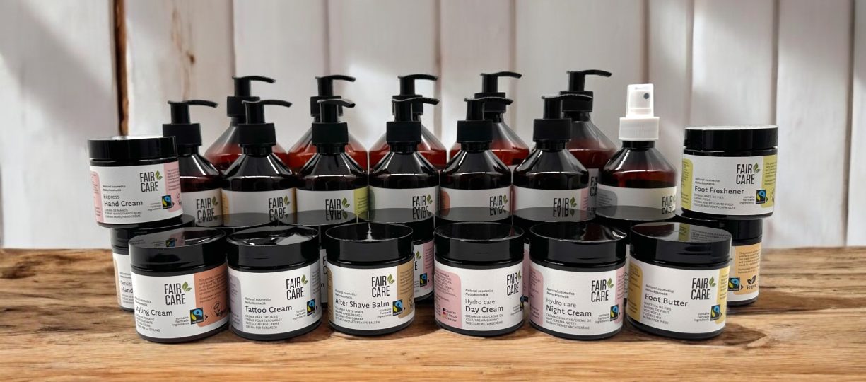 An array of body care products are lined up on a wooden table. Small jars include night cream and foot butter. Large containers with bumps for shampoo, conditioner and shower gel stand behind them.