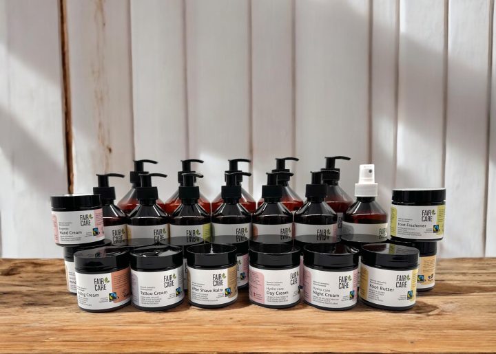 An array of body care products are lined up on a wooden table. Small jars include night cream and foot butter. Large containers with bumps for shampoo, conditioner and shower gel stand behind them.