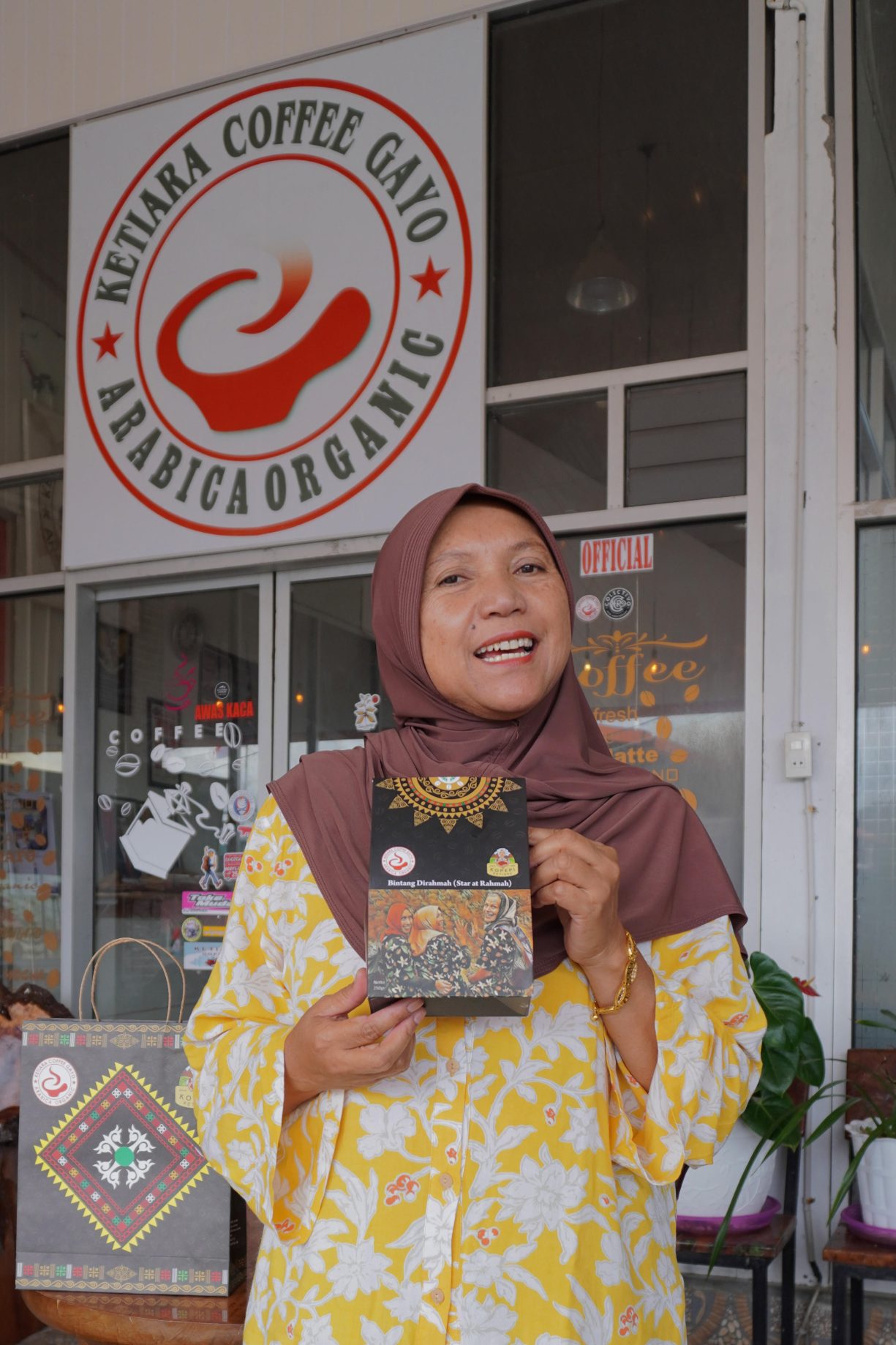 Ibu Rahmah stands holding a bag of coffee in front of a building with glass doors and walls and which has a sign for organic Arabica coffee from Ketiara Coffee Gayo.