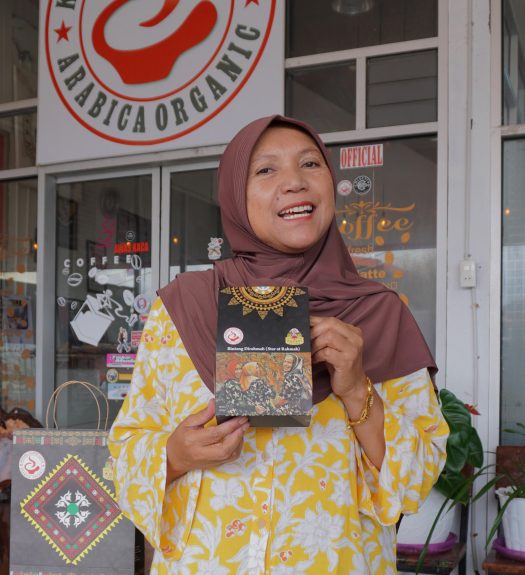 Ibu Rahmah stands holding a bag of coffee in front of a building with glass doors and walls and which has a sign for organic Arabica coffee from Ketiara Coffee Gayo.