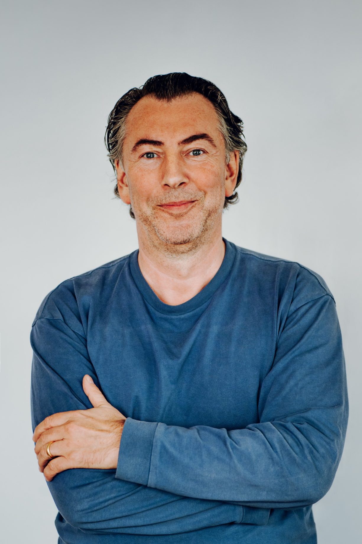 Portrait of Oliver Gothe standing in a long-sleeve blue shirt with his arms gently crossed and smiling with his mouth closed.