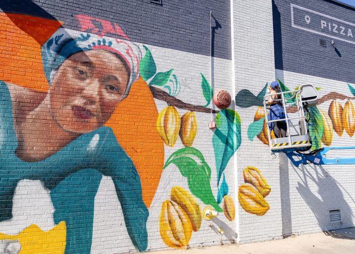 Artist Chelsea Lewinski is raised on a lift to paint mural of cocoa farmer Salamatu Katta.