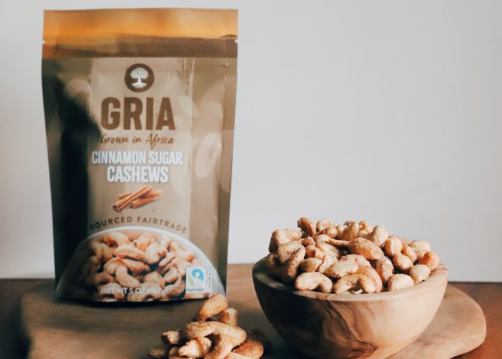 A bag of GRIA’s Fairtrade Cinnamon Sugar Cashews sits atop a wooden board. To the right of the bag is a wooden bowl displaying those cashews. Alongside it is a shallow bowl with cinnamon sugar.