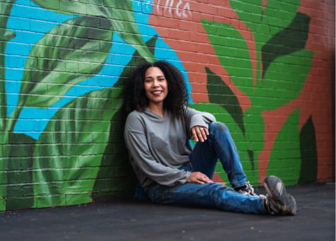 Artist Julia Bottoms smiles as she sits in a relaxed pose against the brick wall where she has created a mural that is rich with foliage.