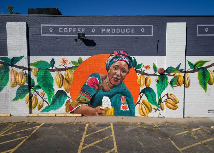 A mural featuring cocoa farmer Salamatu Katta who wears a vibrantly patterened dress and holds an open cocoa pod amid green foliage and ripe, yellow cocoa pods. Above the mural, signage on the storefront reads: 