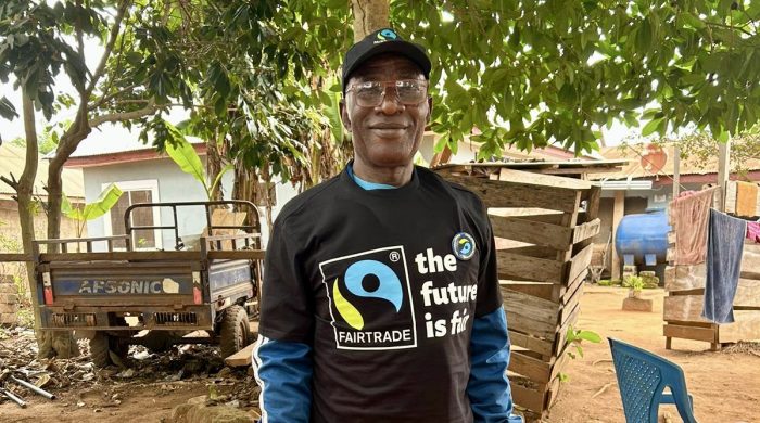 Ghanaian cocoa farmer Daniel Amponsah smiles to the camera as he stands in front of his home. He wears a Fairtrade t-shirt.