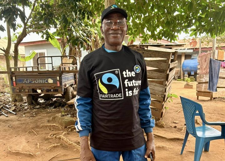 Ghanaian cocoa farmer Daniel Amponsah smiles to the camera as he stands in front of his home. He wears a Fairtrade t-shirt.