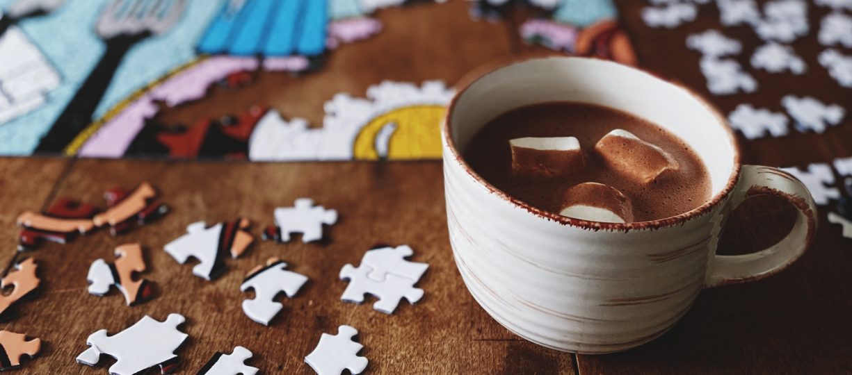 On a wooden table is a work-in-progress jigsaw puzzle and a mug of hot chocolate with marshmallows.