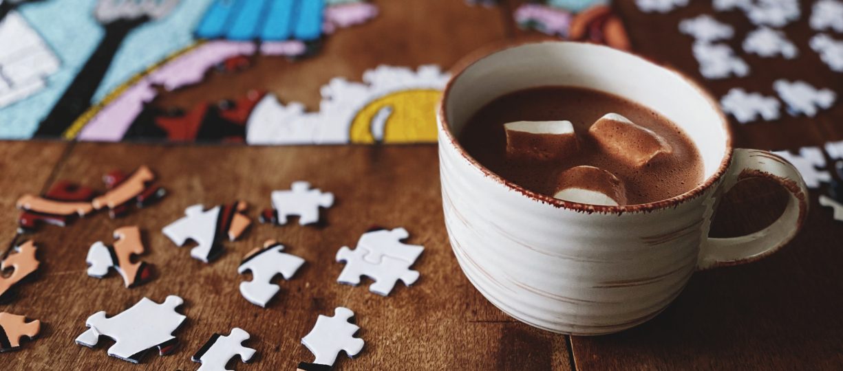 On a wooden table is a work-in-progress jigsaw puzzle and a mug of hot chocolate with marshmallows.