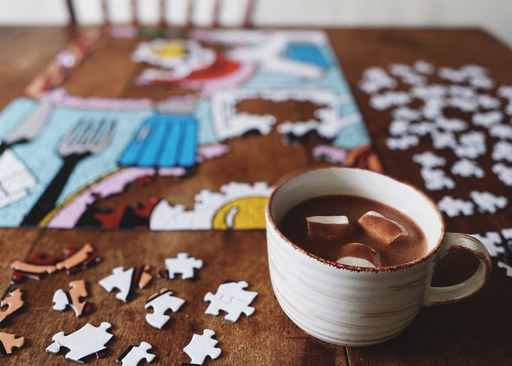 On a wooden table is a work-in-progress jigsaw puzzle and a mug of hot chocolate with marshmallows.