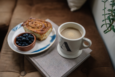 A cozy scene at home where a book lies on a couch and holds a plate of pastry and fruit alongside a hot cup of coffee.