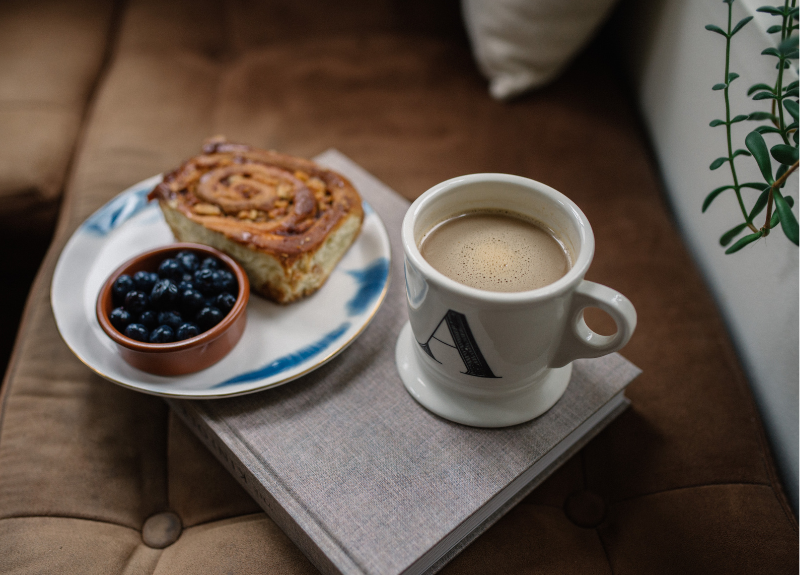 A cozy scene at home where a book lies on a couch and holds a plate of pastry and fruit alongside a hot cup of coffee.