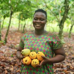 Dora Atiiga holds a pile of five, yellow cocoa pods and smiles as she stands amid her cacao trees.