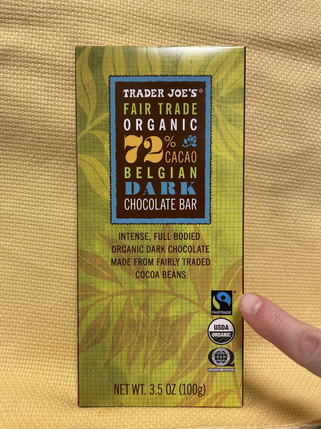 A finger points to the blue, green, and black Fairtrade Mark on the packaging for a Trader Joe's Fair Trade Organic 72% Cacao Dark Chocolate Bar.