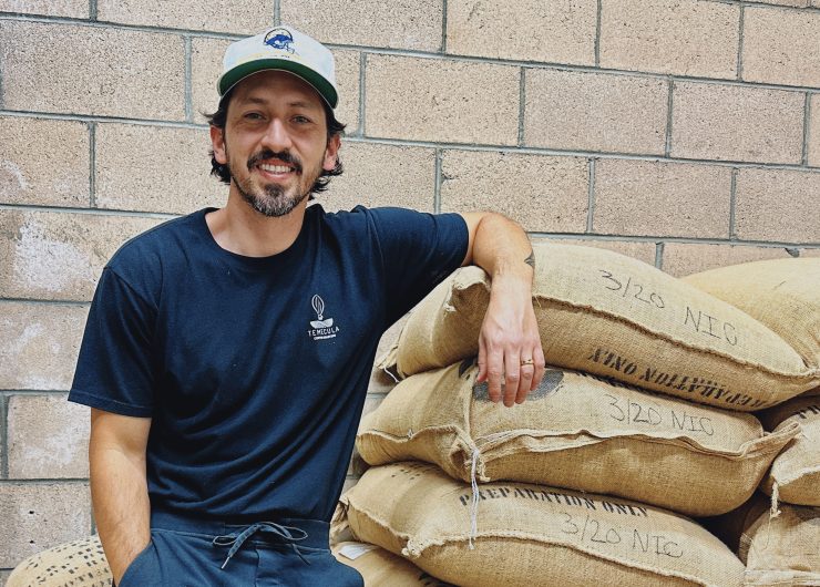 Ed Burga leans against a stack of sacks containing coffee beans. His shirt has the Temecula Coffee Roasters logo on it.