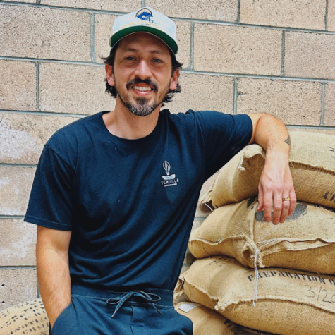 Ed Burga leans against a stack of sacks containing coffee beans. His shirt has the Temecula Coffee Roasters logo on it.